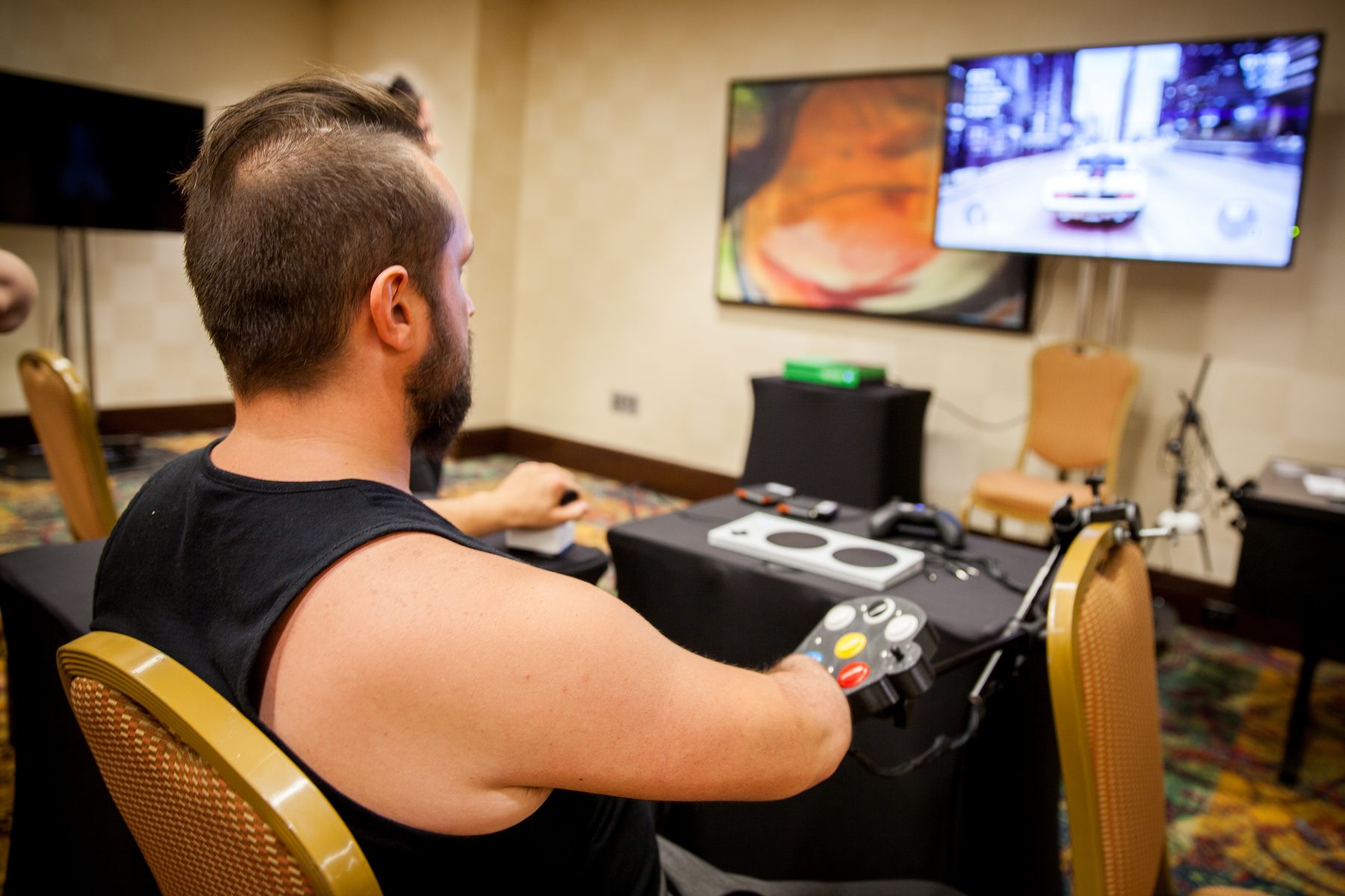 A player seen from behind runs a racing game on a large screen, one hand on a mounted adaptive controller — a console with a joystick and large coloured buttons, held on an articulating arm at the table in front of them.