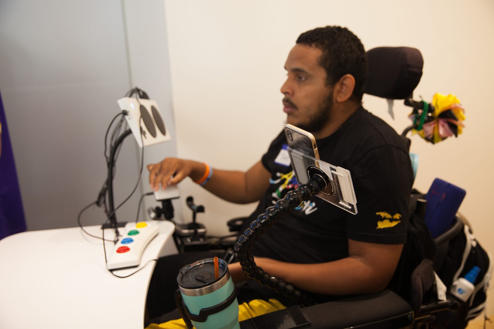 A player using a power wheelchair runs a game with a mounted adaptive-controller rig: a large button box with red, yellow, green, and blue accessible buttons and a phone mounted on a flexible arm, all positioned within easy reach.