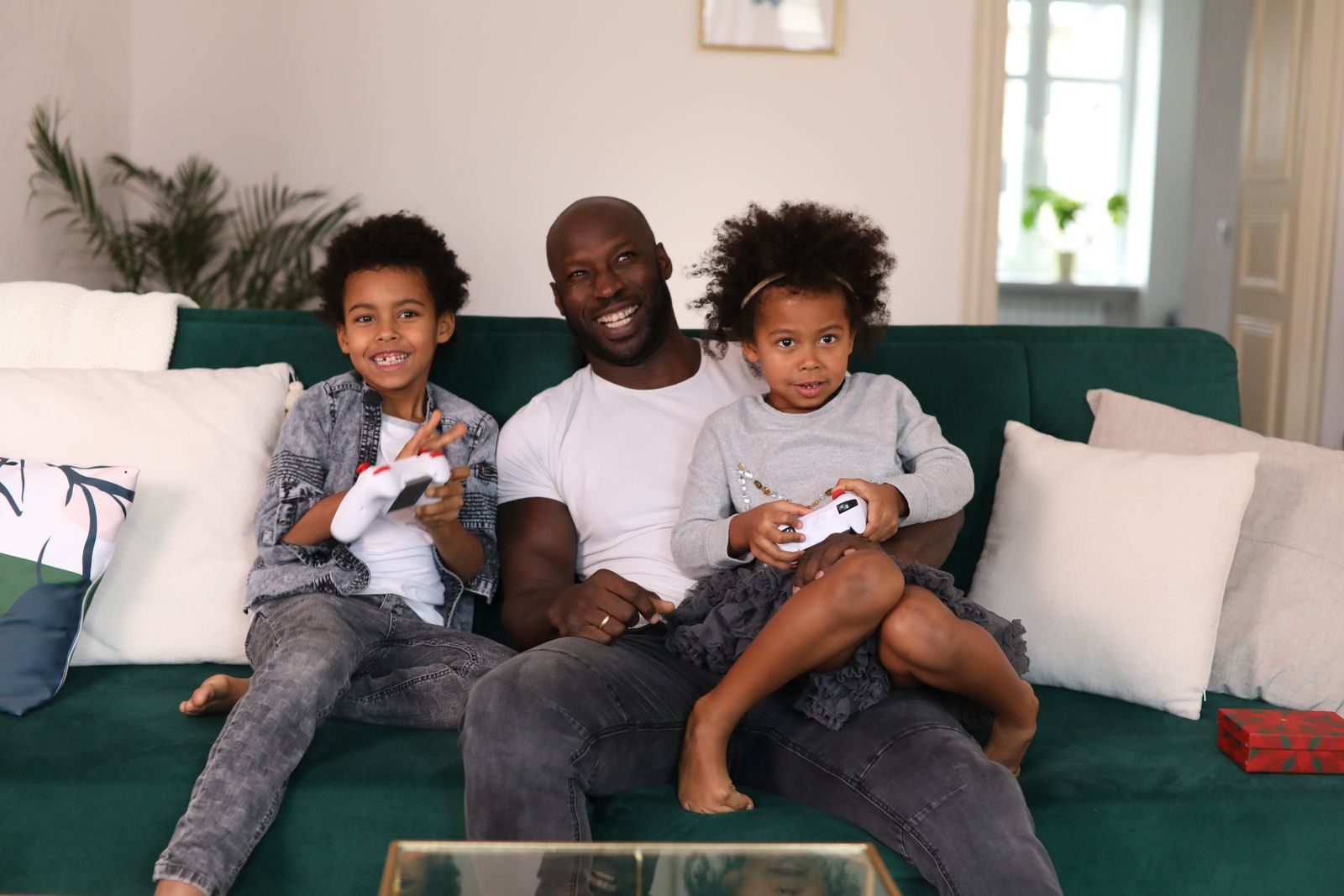 A father and two young children sit together on a couch playing a video game, smiling.