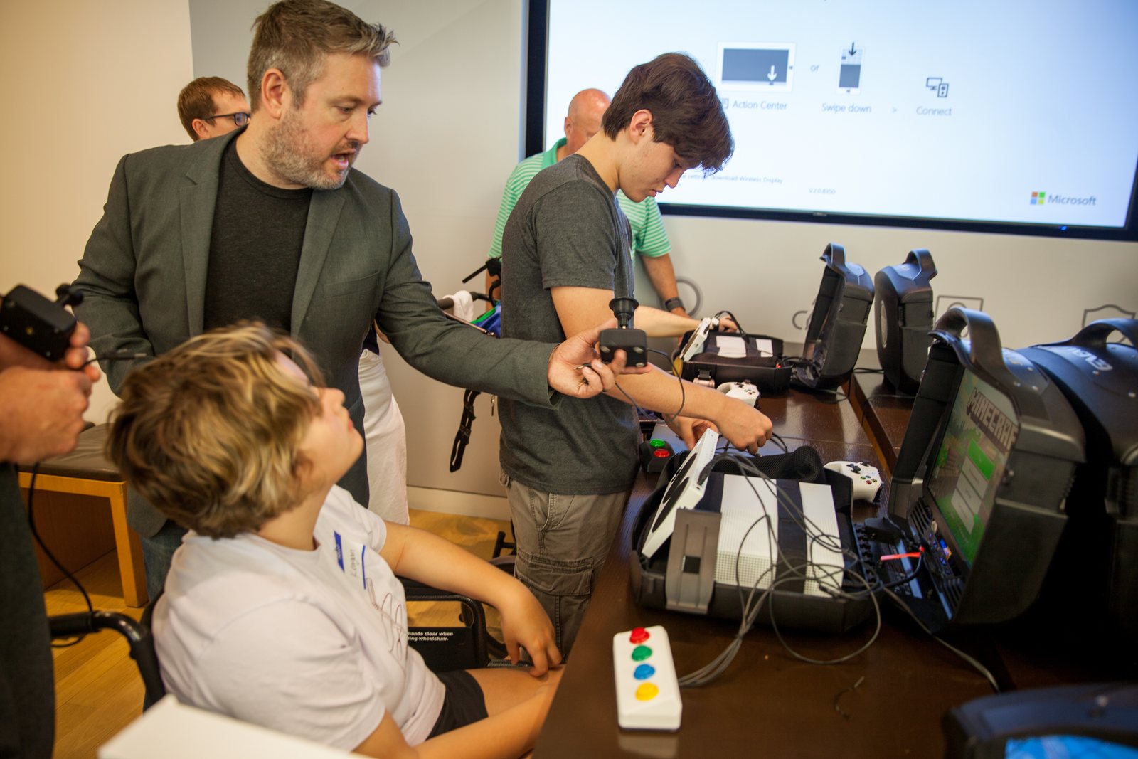 An occupational therapist helps young players set up an adaptive-gaming station — a wheelchair user, an adaptive controller and button box, and Minecraft on the screen.