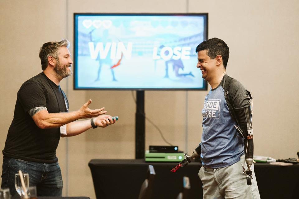 Two players share a laugh during an adaptive-gaming session, a 'Win / Lose' game on the big screen behind them; one plays using bilateral arm prostheses.