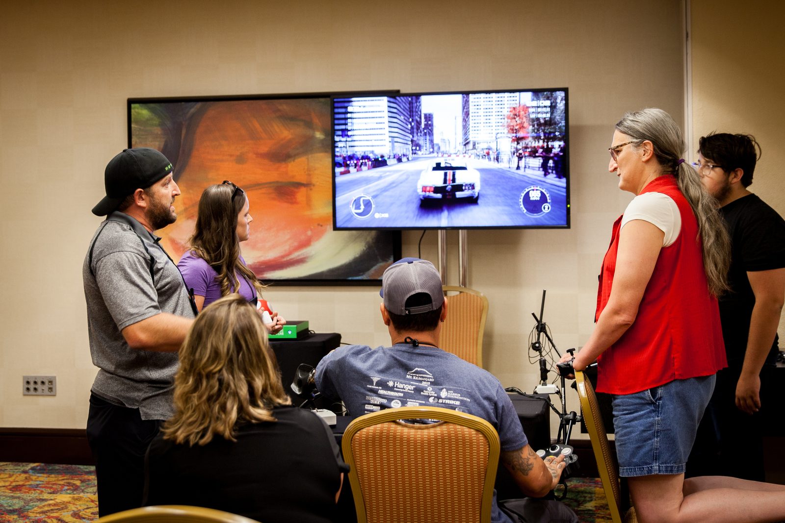 A player uses an adaptive setup to play a racing game on a large screen at a Rehab Arcade session, with others watching.