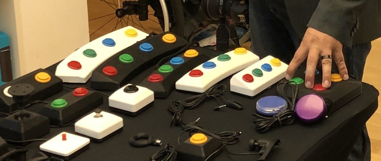 A row of adaptive game controllers on a demo table: white boxes with large red, yellow, green, and blue accessible buttons, alongside dome switches.