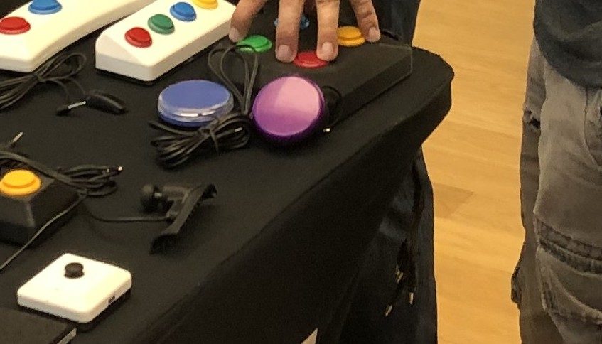 Close-up of large blue and purple dome switches and white boxes with colored accessible buttons on an adaptive gaming demo table.