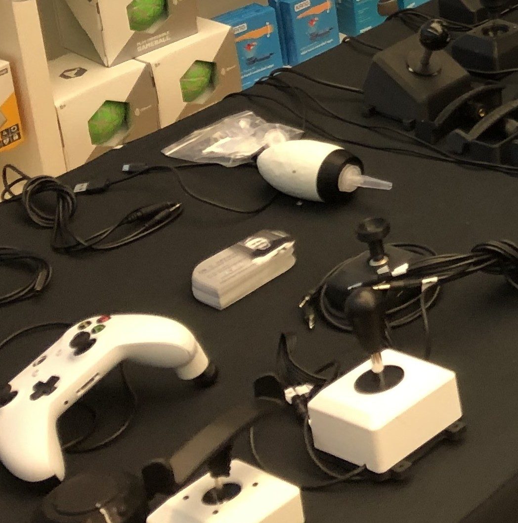 Adaptive joysticks and a controller on an adaptive gaming demo table, with switches and cables.
