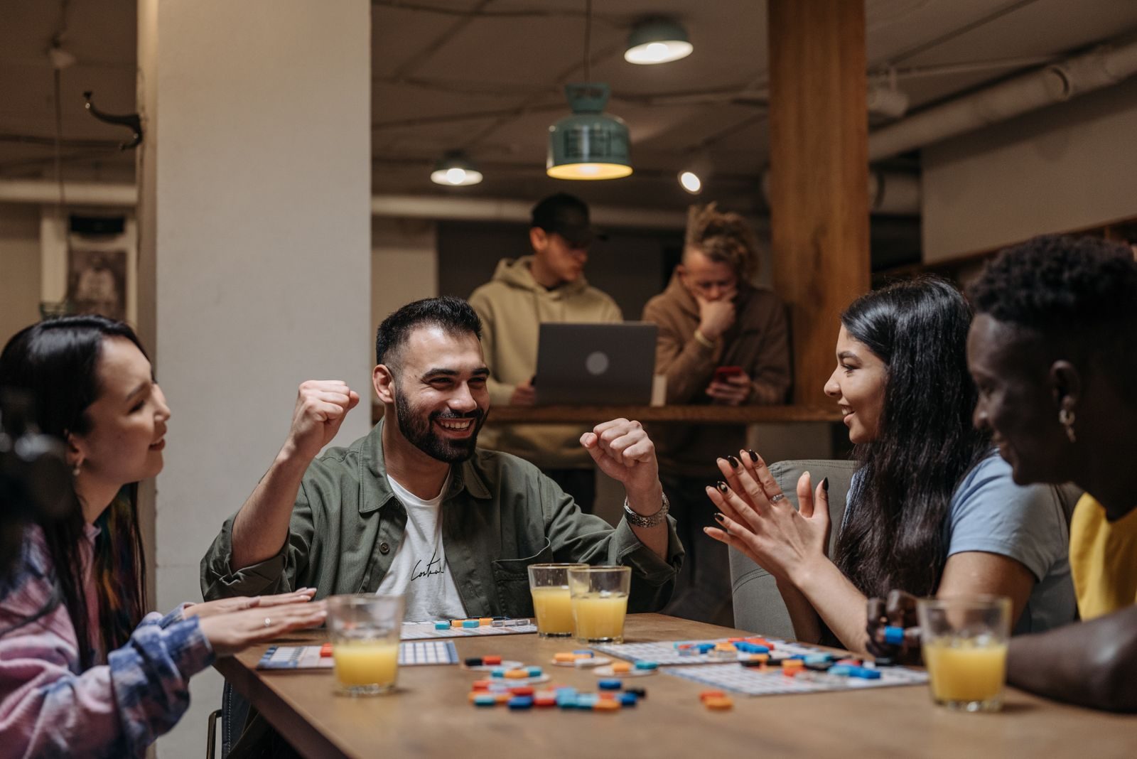 A diverse group of adults seated around a table playing a board game together, smiling and reacting as one player gestures — an engaged, connected small group.