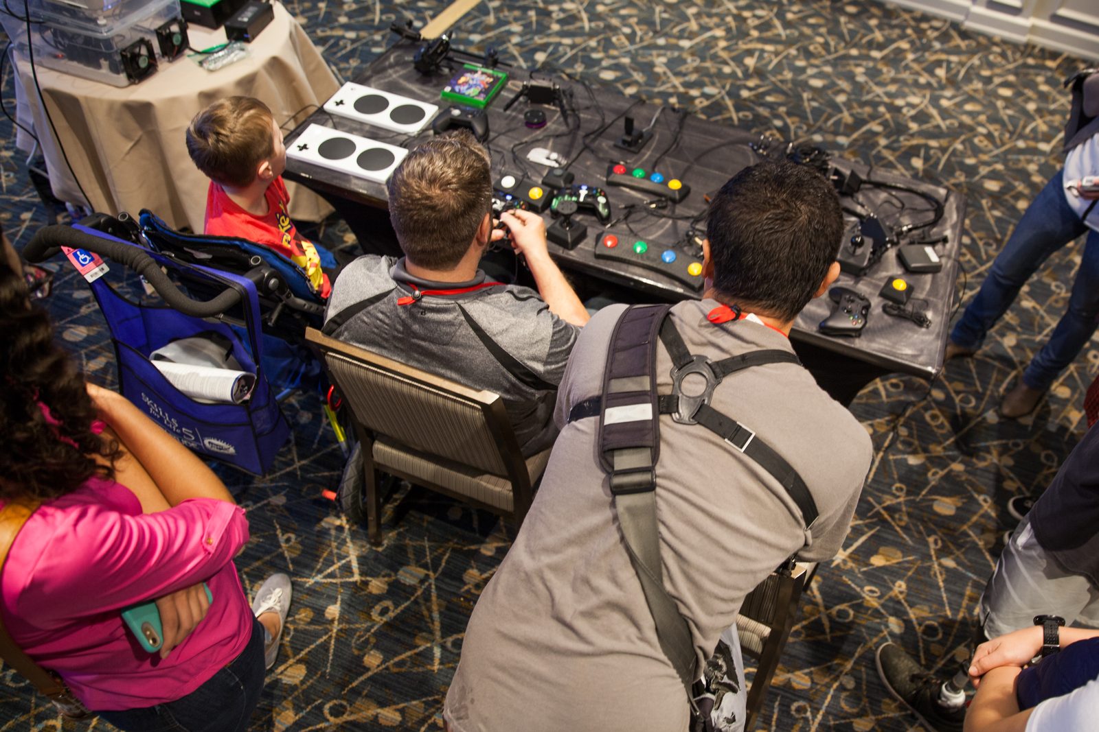 A hands-on table of adaptive game controllers — Xbox Adaptive Controllers, joysticks, and large accessible buttons — where a young player in a wheelchair and helpers try out different setups together.
