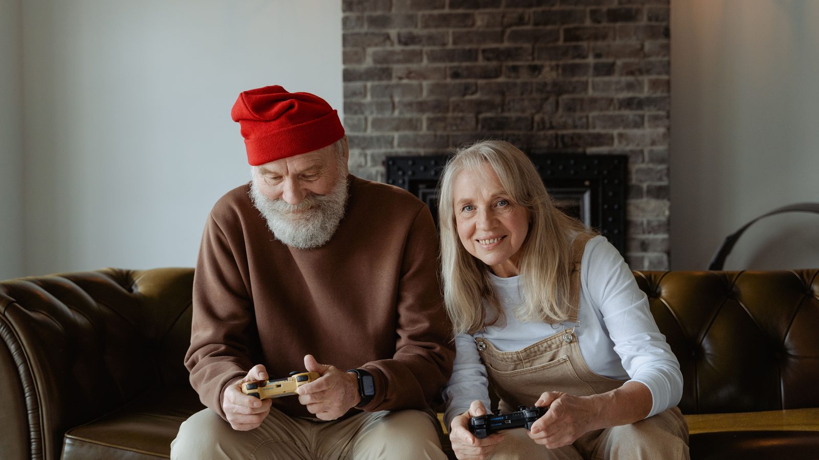 Two older adults sitting together on a sofa, each holding a game controller and playing a video game.